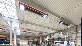 Blick in eine Produktionshalle mit Geräten. An der Decke hängen das multivalente Infrarotsystem zum Heizen.