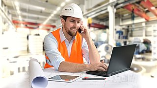 Eine Person in Arbeitsschutzkleidung sitzt an einem Tisch vor einem Laptop. Dabei telefoniert er lächelnd mit seinem Handy.