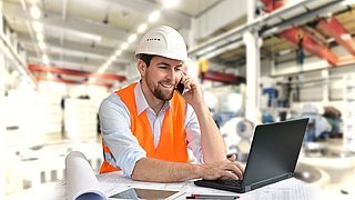 Eine Person in Arbeitsschutzkleidung sitzt an einem Tisch vor einem Laptop. Dabei telefoniert er lächelnd mit seinem Handy.