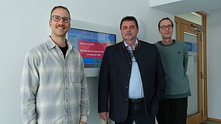 Gruppenaufnahme vor einer digitalen Haustaufel im Flur mit Thomas Koutalidis, Dr. Henri Lüdeke, Frank Bergmann.