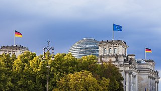 Außenansicht des Reichstaggebäudes. Auf dem Dach wehen die EU- und Deutschlandfahne. Der Vordergrund ist von Bäumen verdeckt.