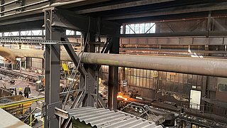 Blick in die Werkshalle des Warmwalzwerks. Im Vordergrund ist ein großes Rohr. Im Hintergrund ist ein Förderband, auf dem eine glutheiße Stahlbramme liegt.