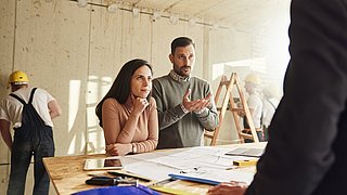 Zwei Personen stehen an einem Tisch und lassen sich von einer gegenüberstehenden Person beraten.