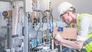 Ein Mann mit Sicherheitsausrüstung notiert etwas auf einem Tablet. Vor ihm ist eine industrielle Anlage. 