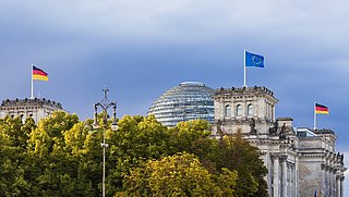Außenansicht des Reichstaggebäudes. Auf dem Dach wehen die EU- und Deutschlandfahne. Der Vordergrund ist von Bäumen verdeckt.