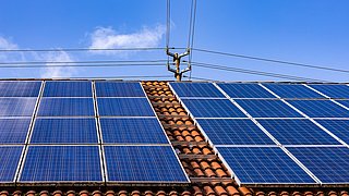 Hausdach mit Photovoltaik-Anlage. Dahinter ragt eine Strom-Oberleitung.