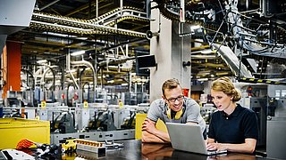 Zwei Personen stehen an einem Tisch in einer Werkshalle und schauen auf einen Laptop