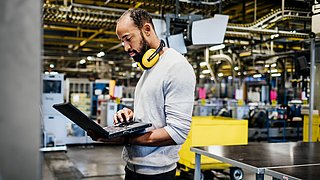 Eine Person steht in einer Werkshalle. Sie hat einen Laptop in der Hand und schaut auf den Bildschirm.