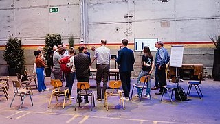 Workshopszene, alles stehen im Kreis um eine White Board und reden miteinander. 