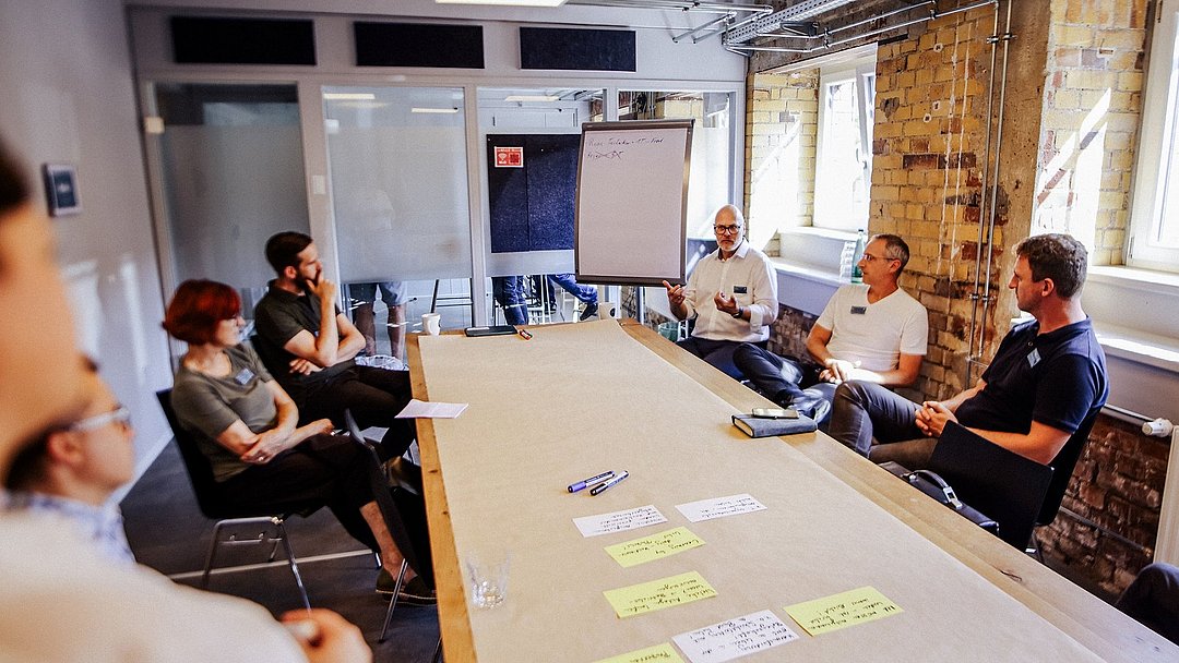 Momentaufnahme des Business Dialogs. Meherere Personen sitzen um einen Tisch herum und sprechen miteinander.