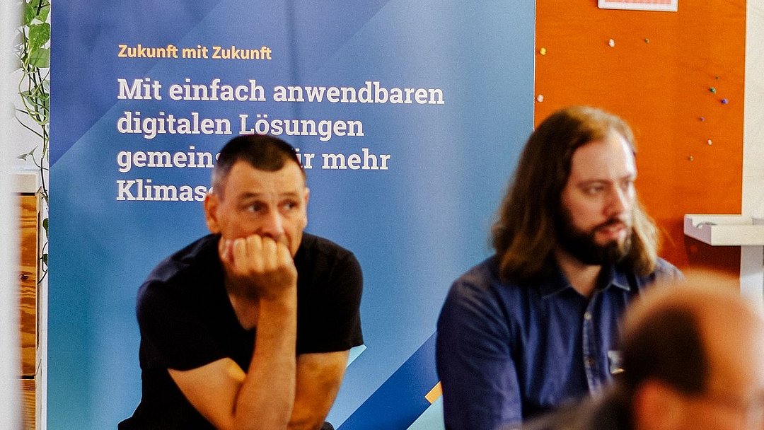 Momentaufnahme des Business Dialogs. Drei Personen sitzen auf Hockern und hören einem Vortrag zu. 