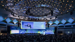 Der Saal des BCC in Berlin. Die Zuschauenden sitzen auf Stühlen und blicken auf die Bühne, auf der eine Person eine Rede hält.