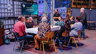 Workshopszene,es sitzen mehrere Menschen um einen Tisch herum und eine Person steht und erzählt etwas. 