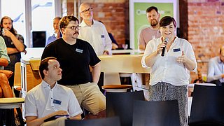 Momentaufnahme des Business Dialogs. Eine Person steht und spricht in ein Mikrofon. Weitere Personen sitzen daneben und hören zu.