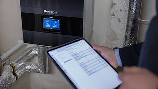 Eine Person hält ein Tablet in der Hand. Dahinter ist die Warmwasseraufbereitung.