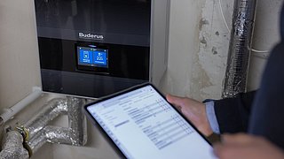Eine Person hält ein Tablet in der Hand. Dahinter ist die Warmwasseraufbereitung.