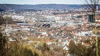 Stadtansicht mit dicht bebautem Innenstadtbereich, Grünflächen und umliegenden Wohn- und Gewerbegebieten. Im Vordergrund sind Gebäude und Straßen deutlich erkennbar, im Hintergrund verläuft der Stadtrand mit Übergang ins Umland.