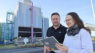 Zwei Personen schauen auf ein Tablet. Im Hintergrund ist das Rückkühlwerk des Unternehmes Infraserv Hoechst.
