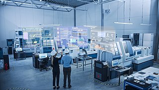 Zwei Personen stehen in einer futuristischen Werkshalle und nehmen ein Foto der Maschinen mit einem Tablet auf.