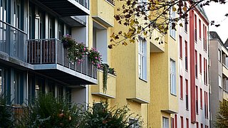 Vorderseiten von verschiedenen Mehrfamilienhäusern. Ganz vorn eine moderne Fassade mit Balkon, daneben ein gelbes und rotes Gebäude.
