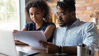 Zwei Personen sitzen an einem Tisch und schauen auf ihre Unterlagen. Davor steht ein Laptop.