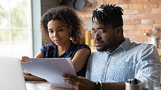 Zwei Personen sitzen an einem Tisch und schauen auf ihre Unterlagen. Davor steht ein Laptop.