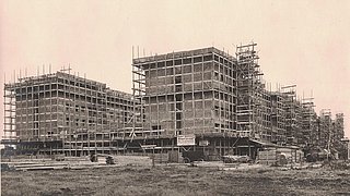 Historische Aufnahme des Baus eines Mehrfamilienhauses. Der Rohbau des Gebäudes ist mit Holzgerüsten verkleidet.