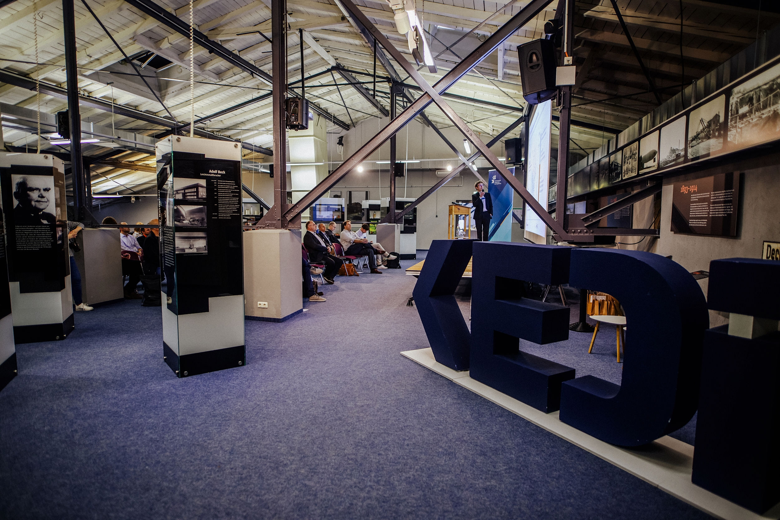 Blick in den Veranstaltungsaal. Im Vordergrund steht ein großes KEDi Logo. Dahinter ist die Bühne und die Teilnehmenden.